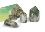 Lodolite Garden Quartz obelisk tower crystal from GemstoneGO with clear and smoky inclusions, standing upright on a white background.